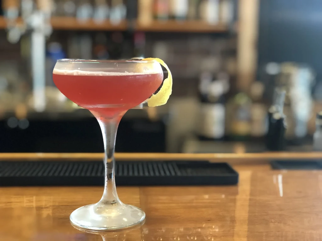 Red cocktail in coupe glass with foam, citrus peel, on bar.