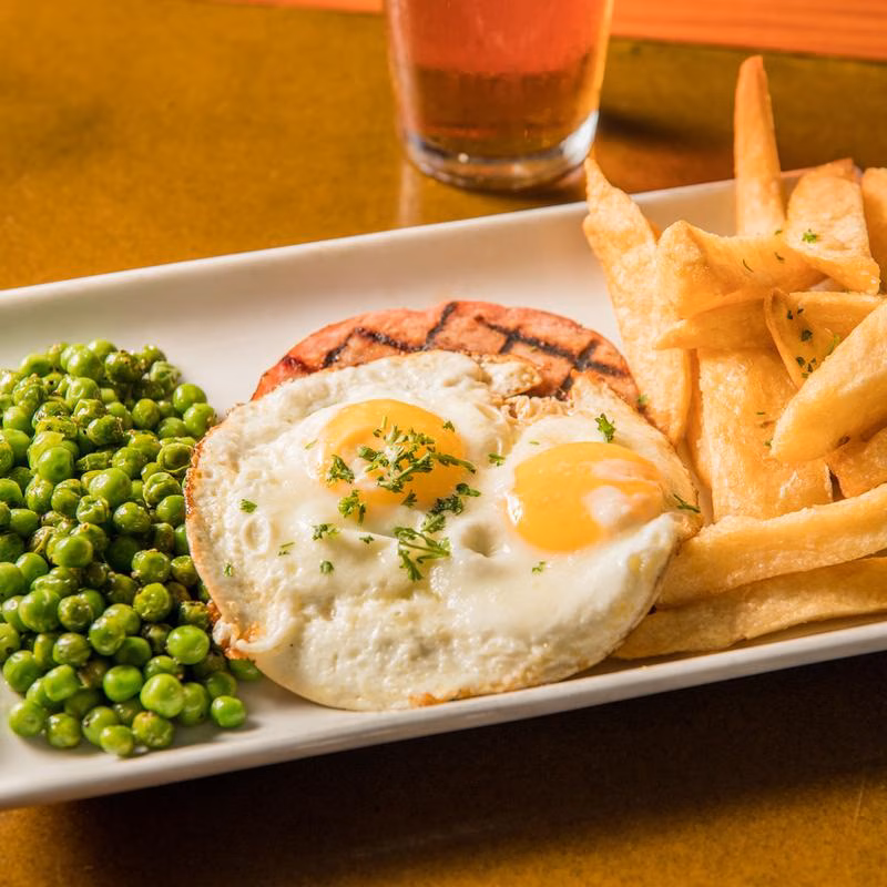 Fried eggs, grilled ham, peas, and fries on a white plate.