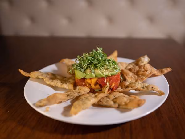 Red and yellow diced food, avocado, microgreens, and crispy chips.