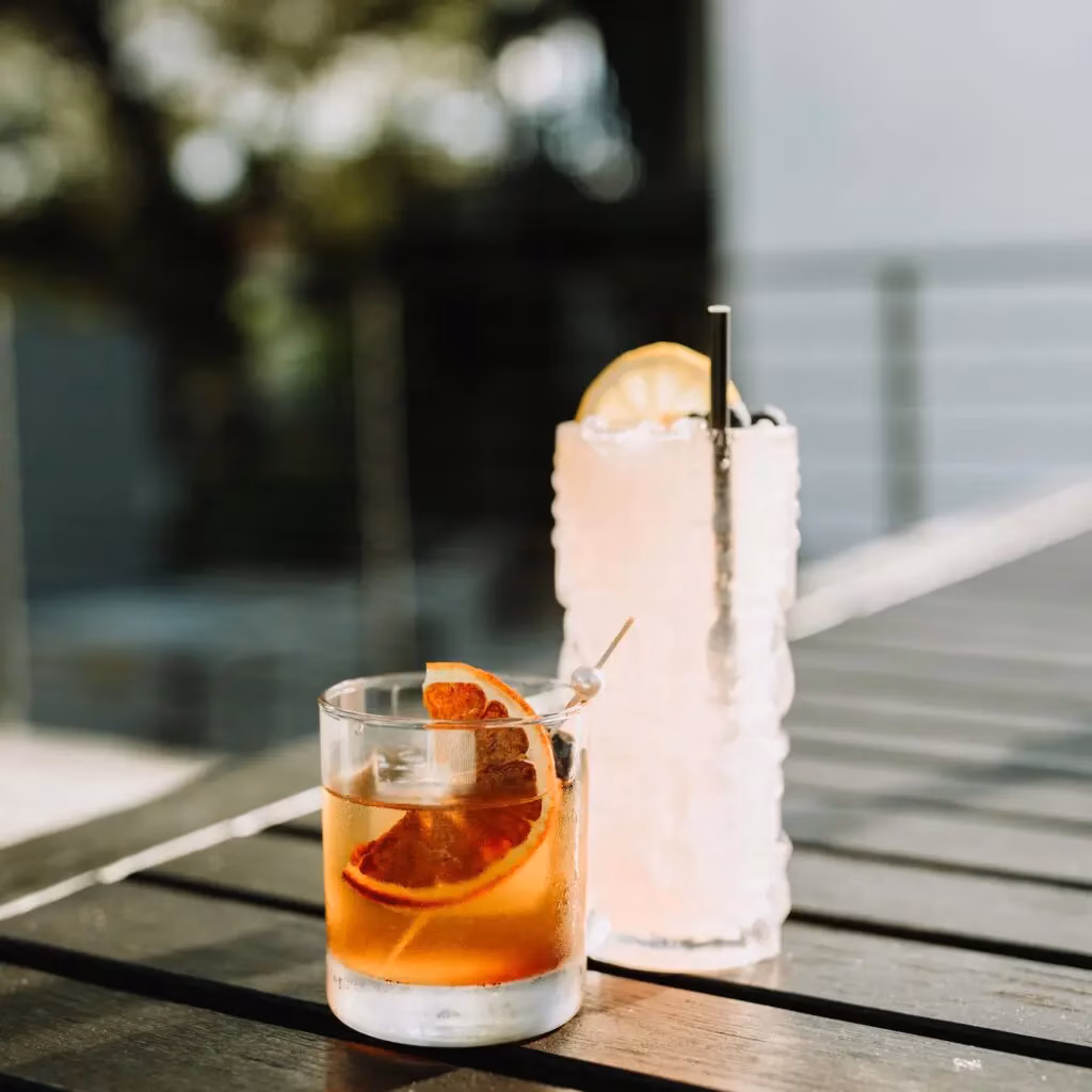 Two cocktails with fruit garnishes on a dark wooden outdoor table.