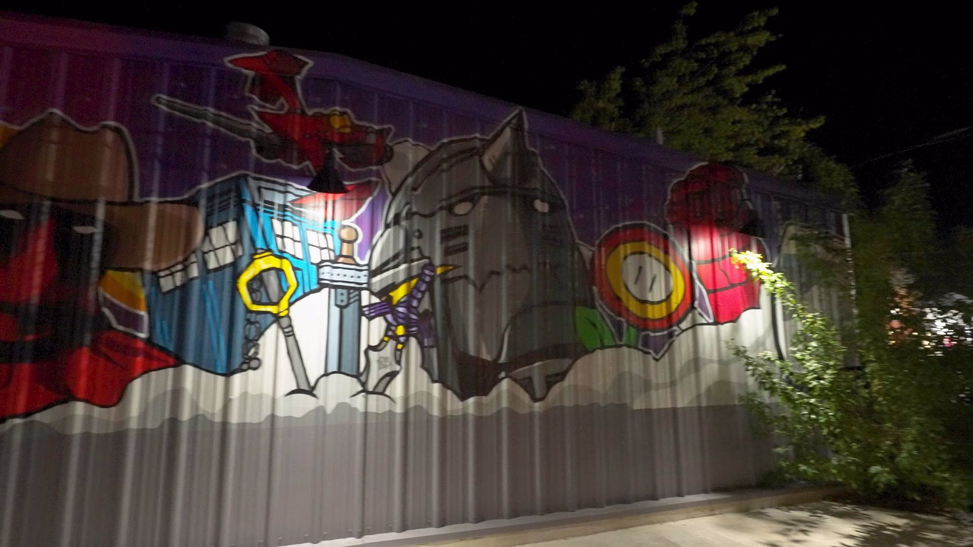 Colorful pop culture mural on corrugated metal wall illuminated at night.