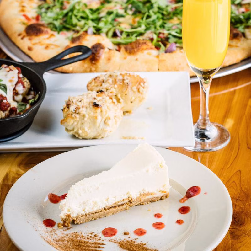 Cheesecake slice, pizza, small skillet, bread rolls, and mimosa on table.