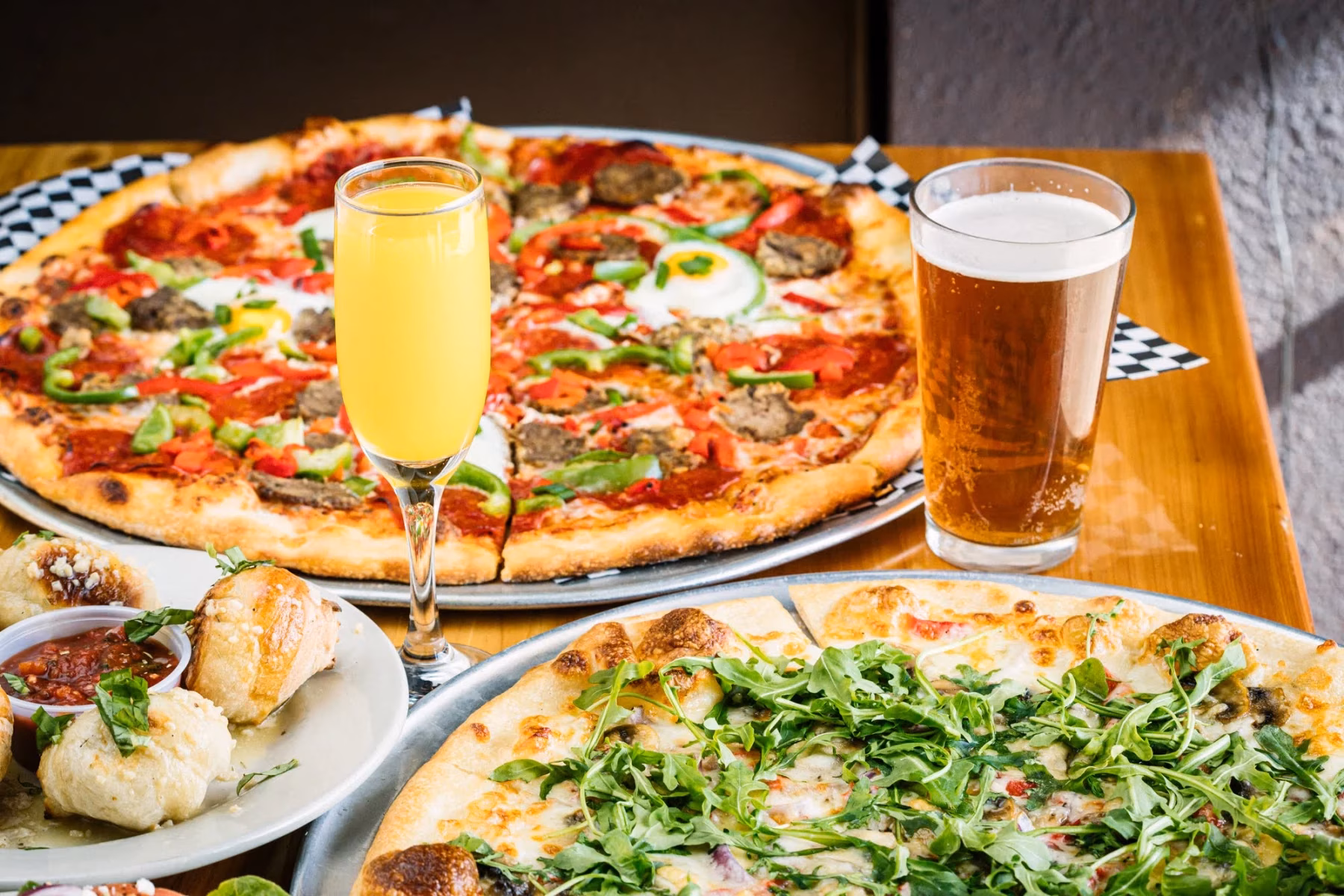 Two pizzas, one with egg, an orange drink, beer, and bread.