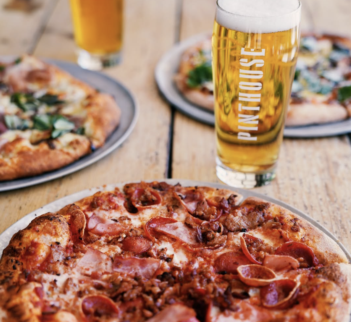 Pizzas and a Pinthouse beer glass on a wooden table.