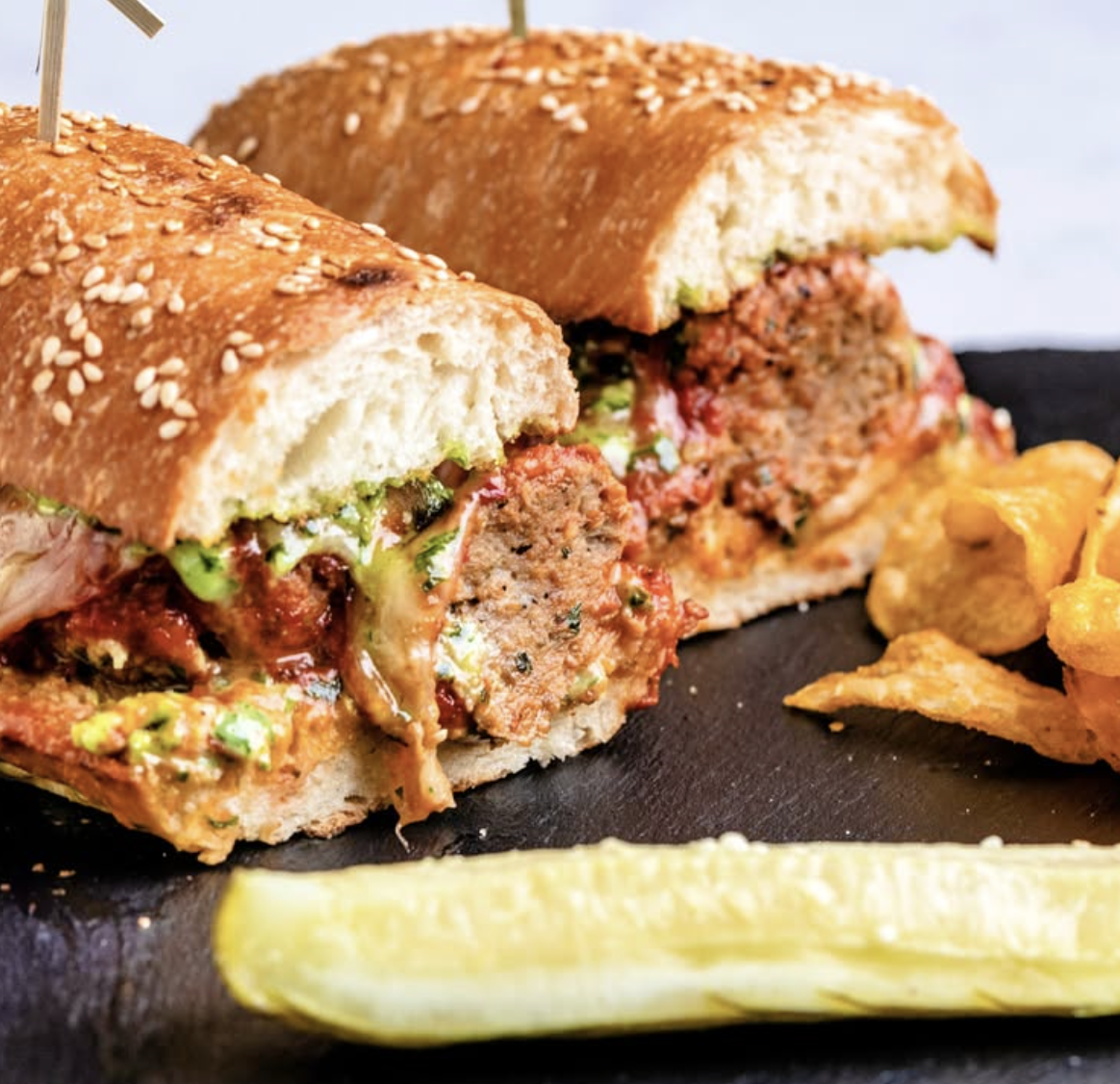 Two meatball subs with sauces, chips, and a pickle.
