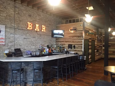 Empty bar interior with stools, bottles, stone wall, and "BAR" sign.