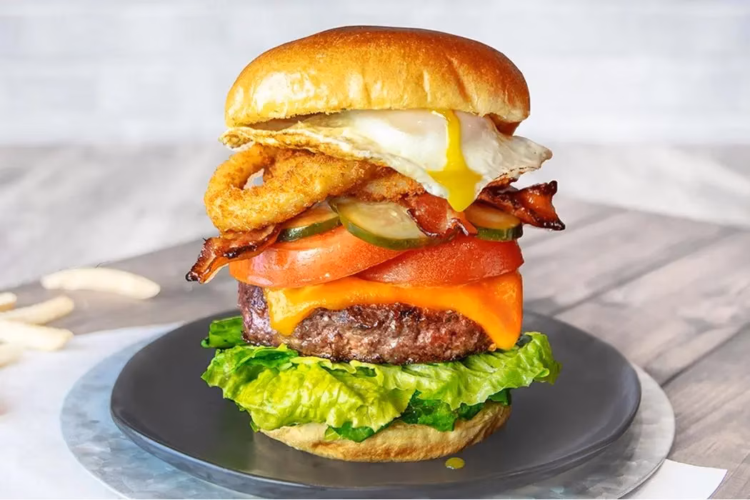 Stacked burger with egg, bacon, onion rings, cheese, lettuce, and tomato.