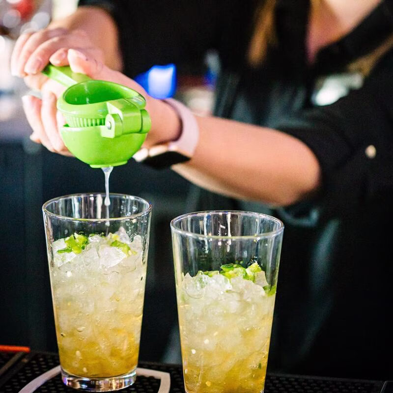 Hands squeezing lime into glasses with ice and green pieces.