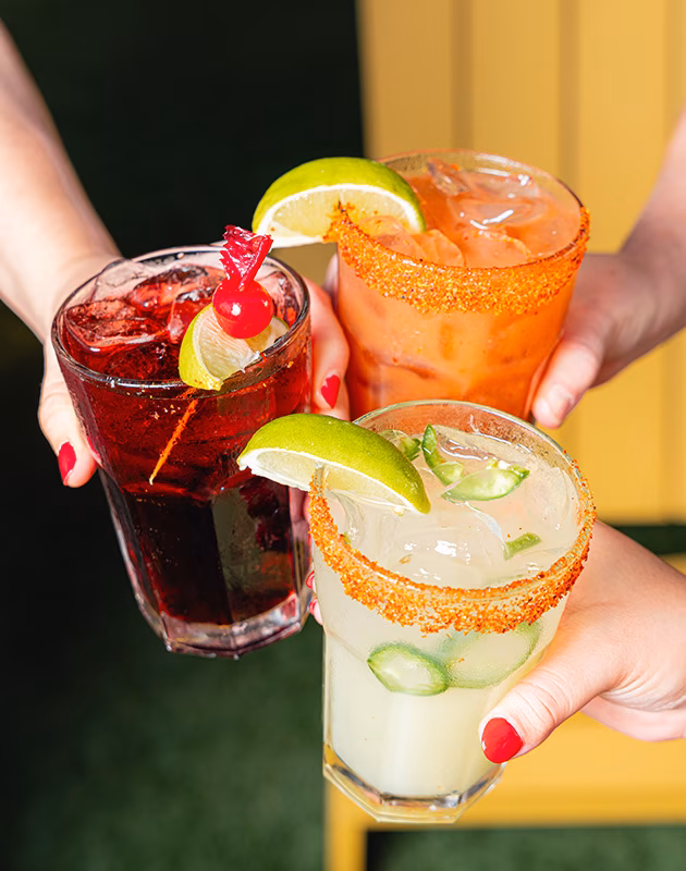 Three hands holding different colorful cocktails, two with spiced rims.