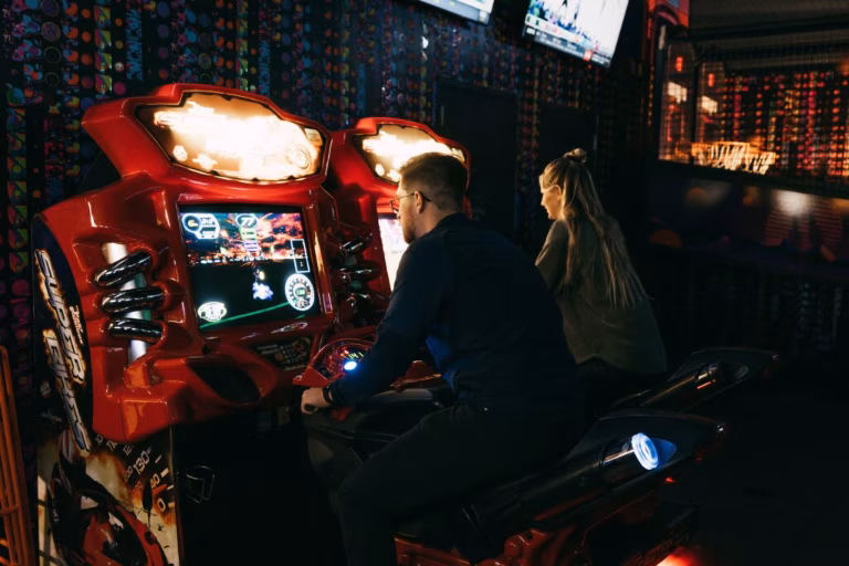 Man plays motorcycle arcade game, woman sits on adjacent machine.