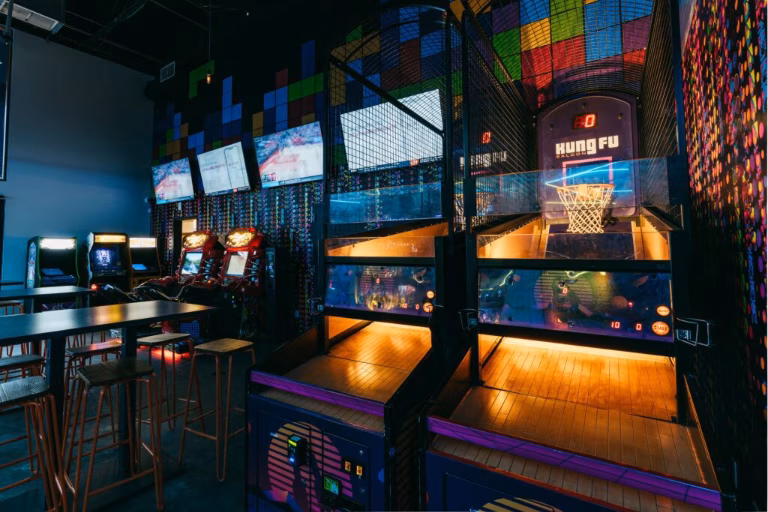 Indoor arcade with basketball games, classic machines, tables, and neon lighting.