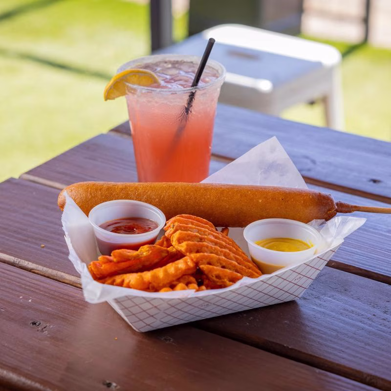 Corn dog, waffle fries, sauces, and iced drink on a wooden table.