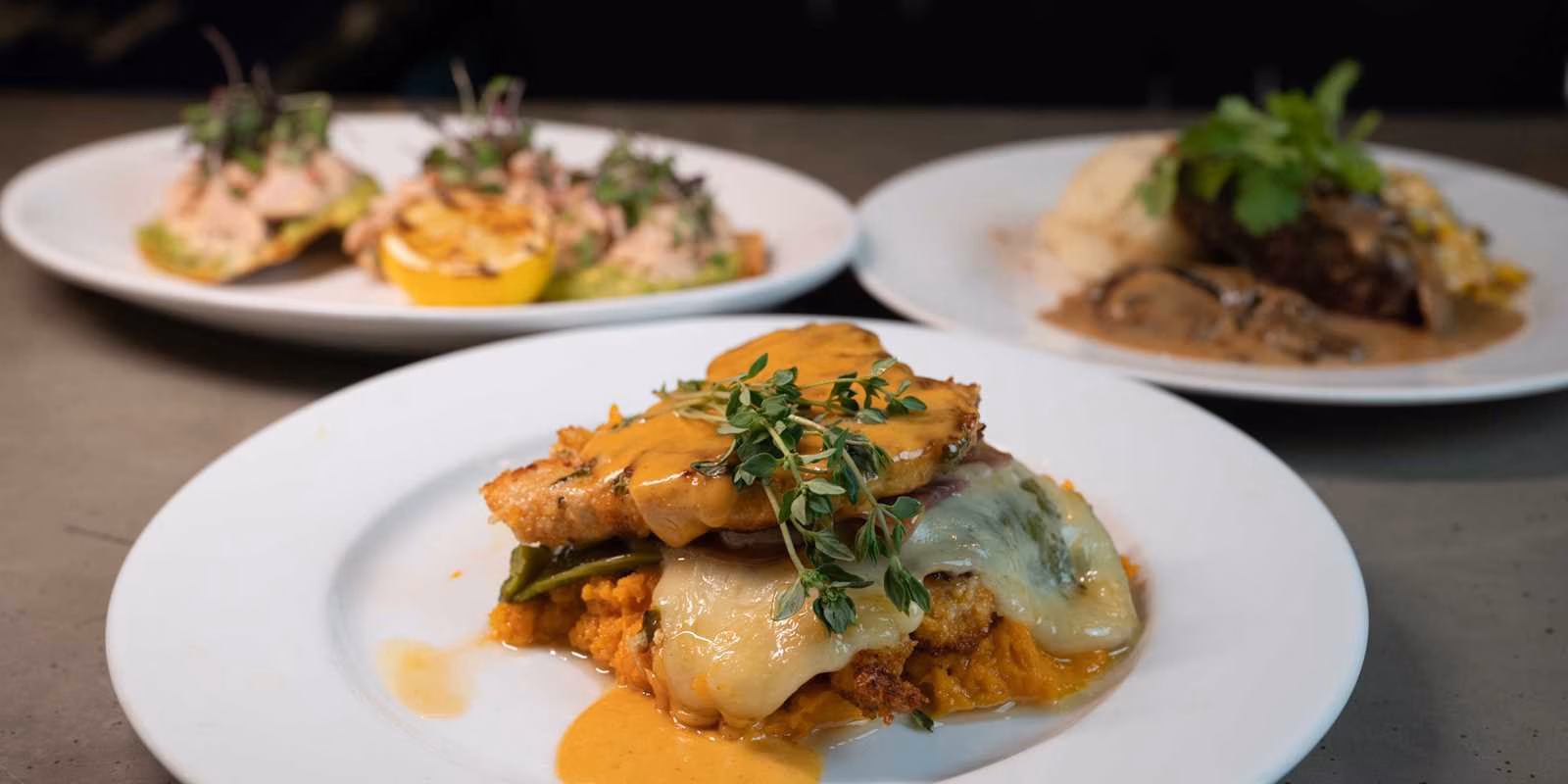 Front dish: seared protein with cheese, sauce, herbs; two other plates.