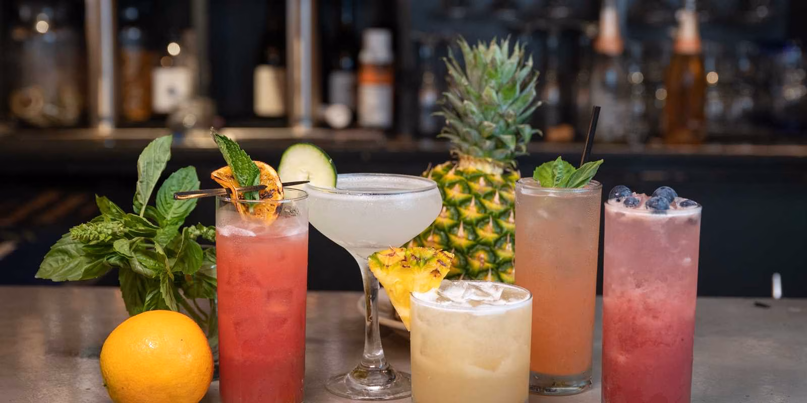 Five colorful cocktails with various fruit and herb garnishes on a bar.