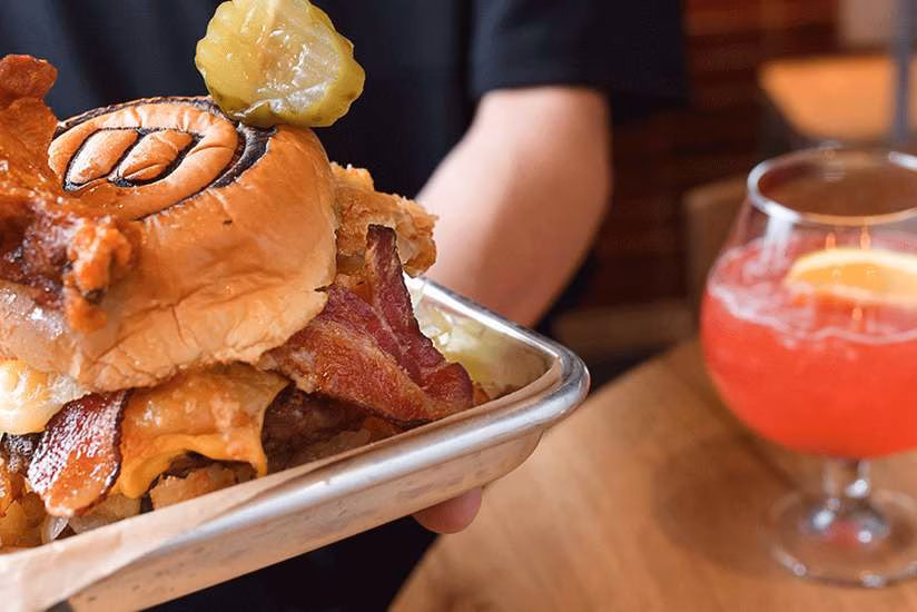 Person holding a large burger with a pickle, bacon, and red drink.