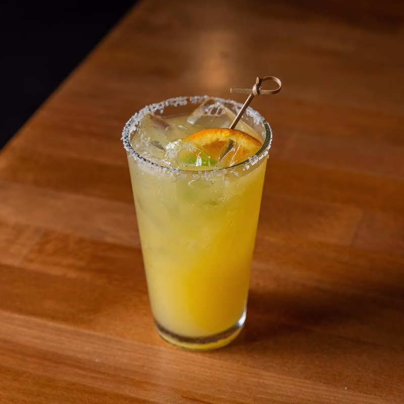 Yellow iced drink in a salt-rimmed glass with orange garnish.