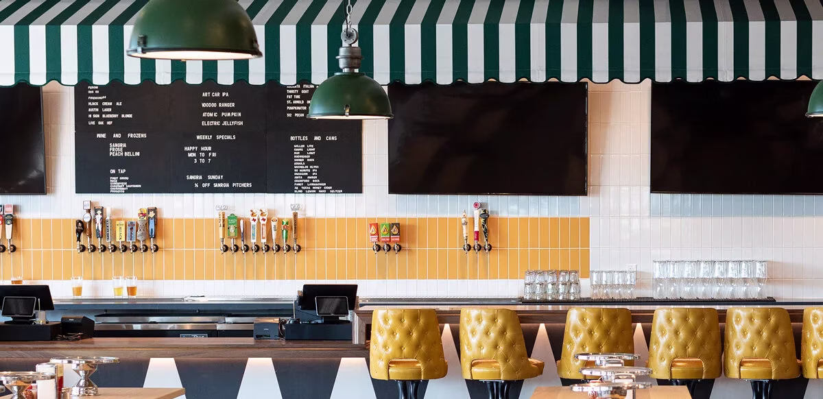 Modern bar interior with green-striped awning, menu boards, beer taps, and yellow stools.