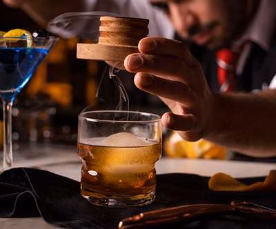 Bartender smokes an amber cocktail with a wooden device over ice.