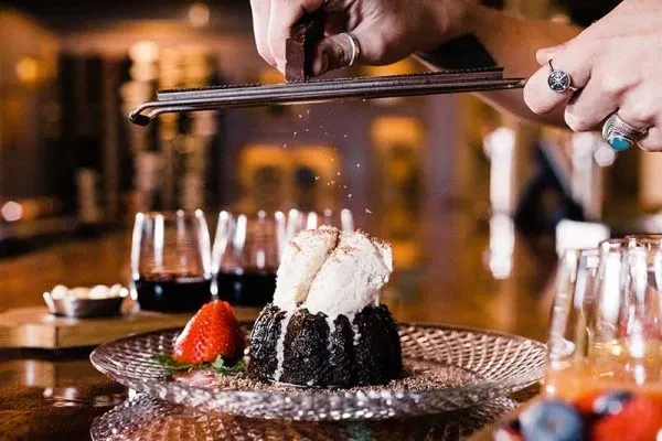 Hands grate chocolate over molten lava cake, ice cream, and strawberry.