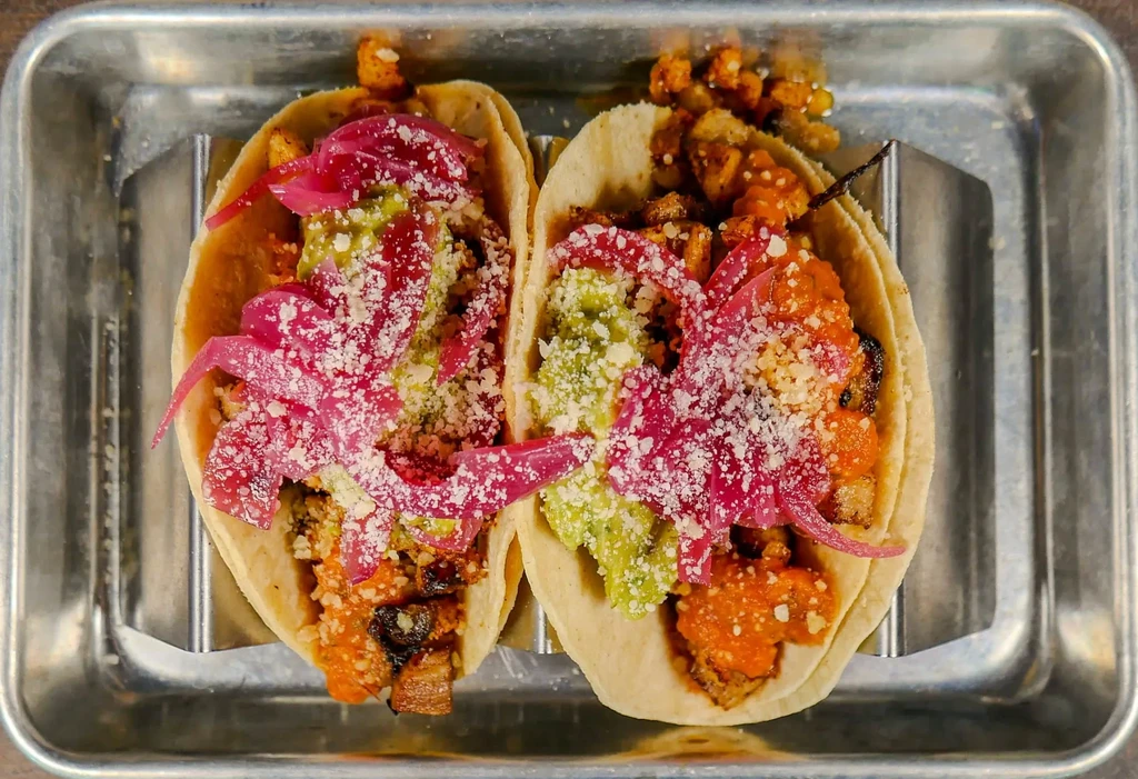 Two tacos with guacamole, pickled onions, cheese, and protein in a holder.