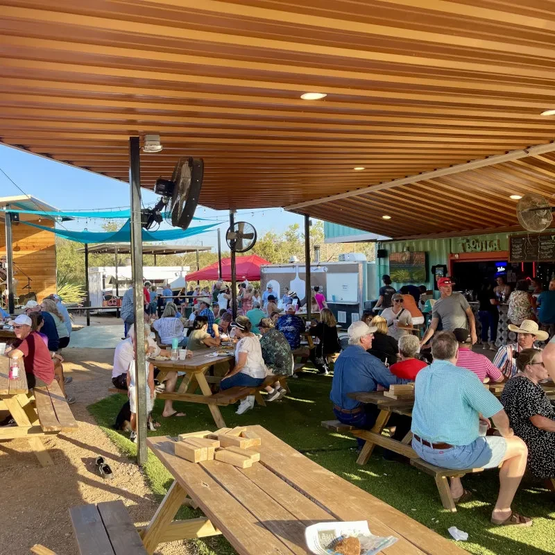 Lively outdoor restaurant with picnic tables, shade canopy, and container vendors.