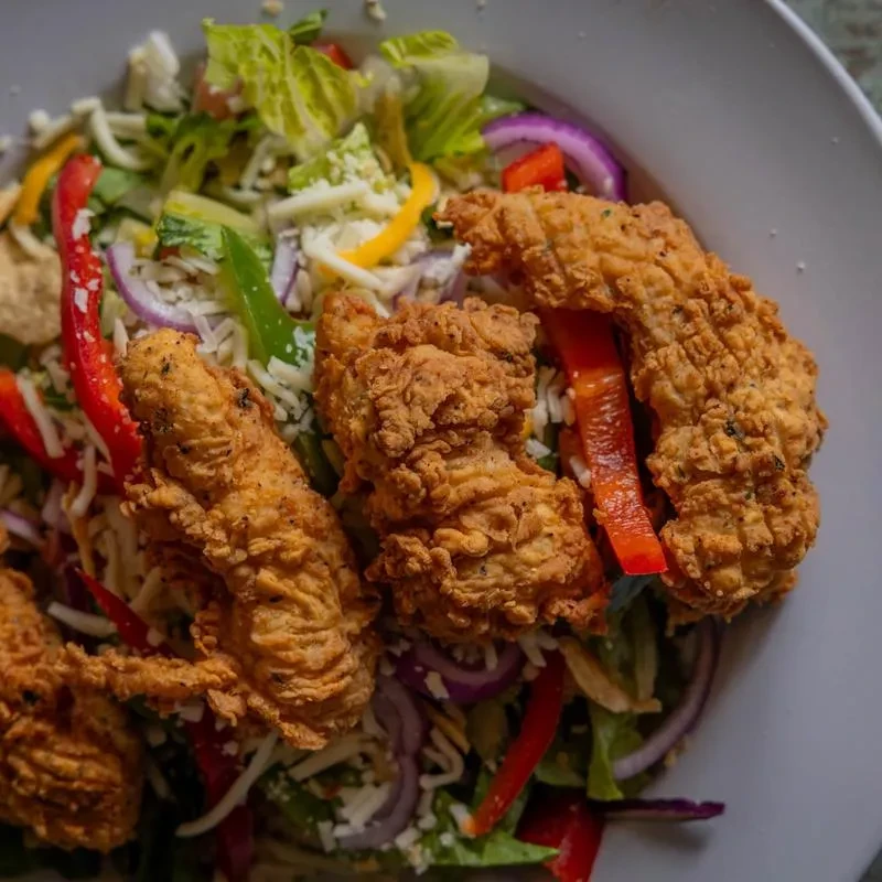 Crispy fried chicken salad with greens, peppers, cheese in white bowl.