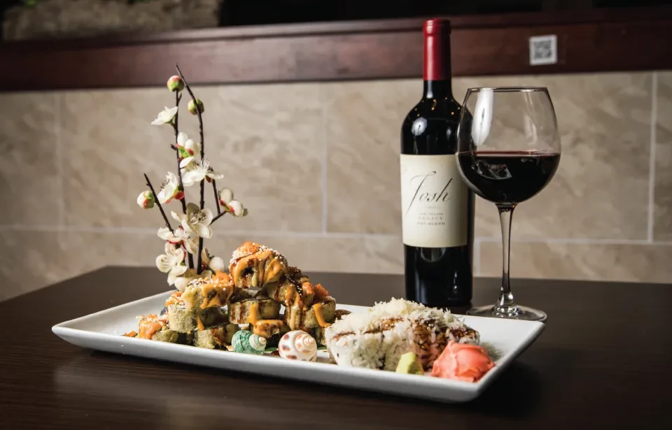 Sushi plate with flowers, red wine bottle and glass on wood table.
