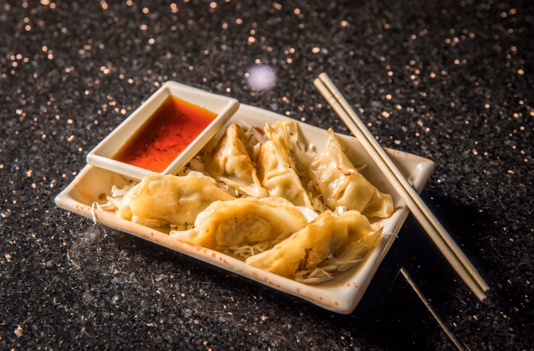 Dumplings with red dipping sauce and chopsticks on a dark countertop.