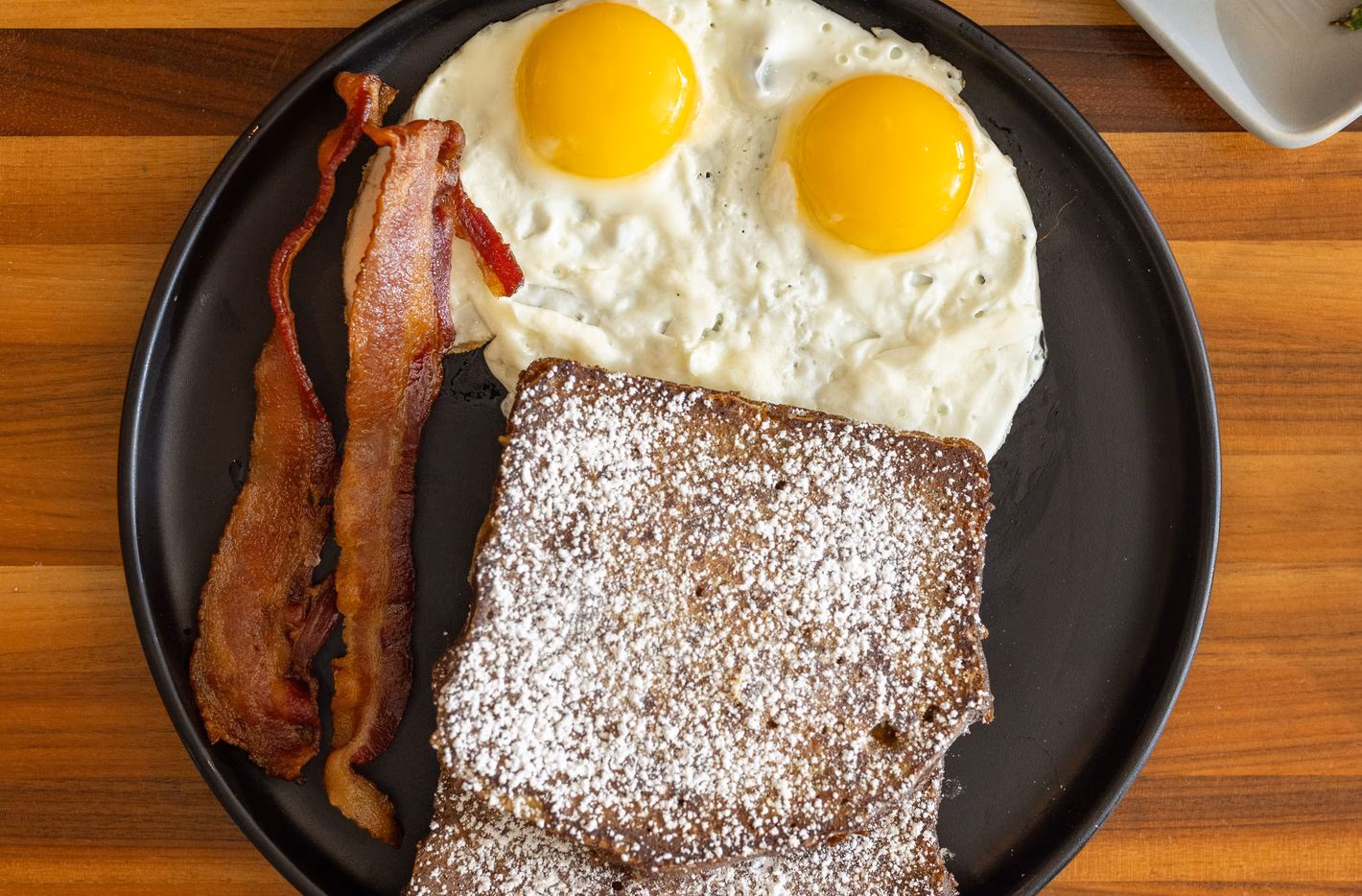 French toast with powdered sugar, two fried eggs, and bacon.