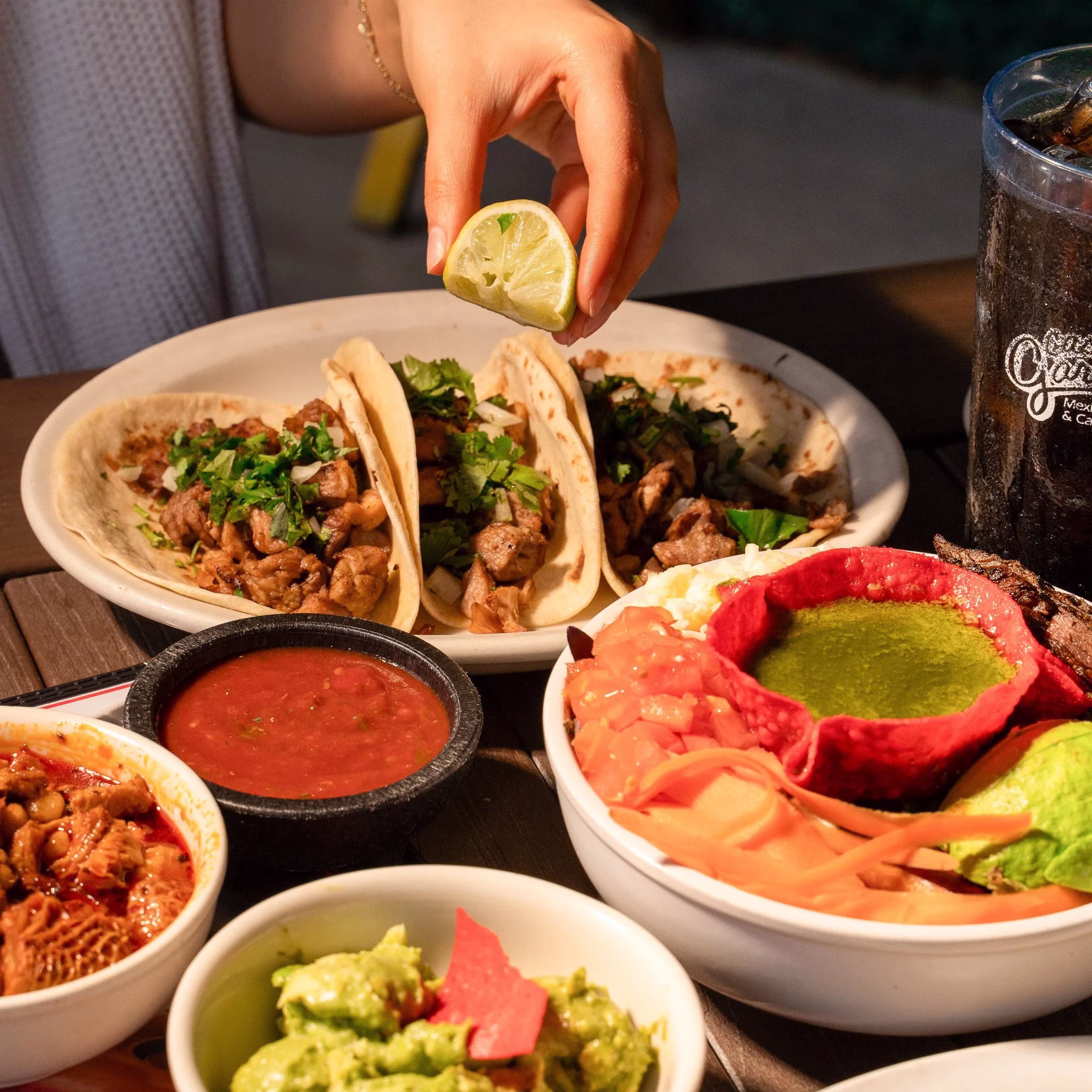 Hand squeezing lime over tacos, surrounded by various Mexican foods.