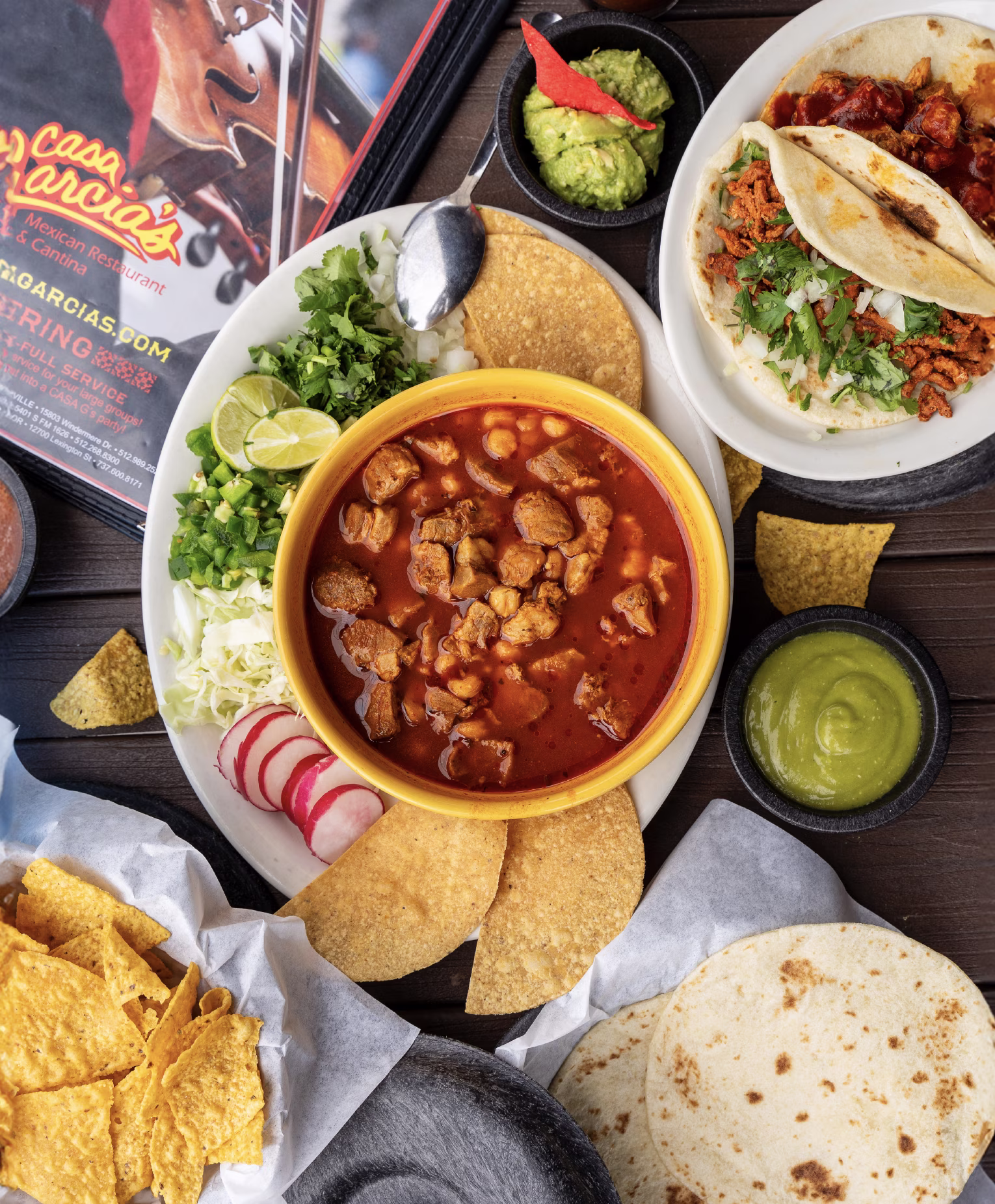 Overhead view of a Mexican meal: red stew, tacos, chips, guacamole, salsa.