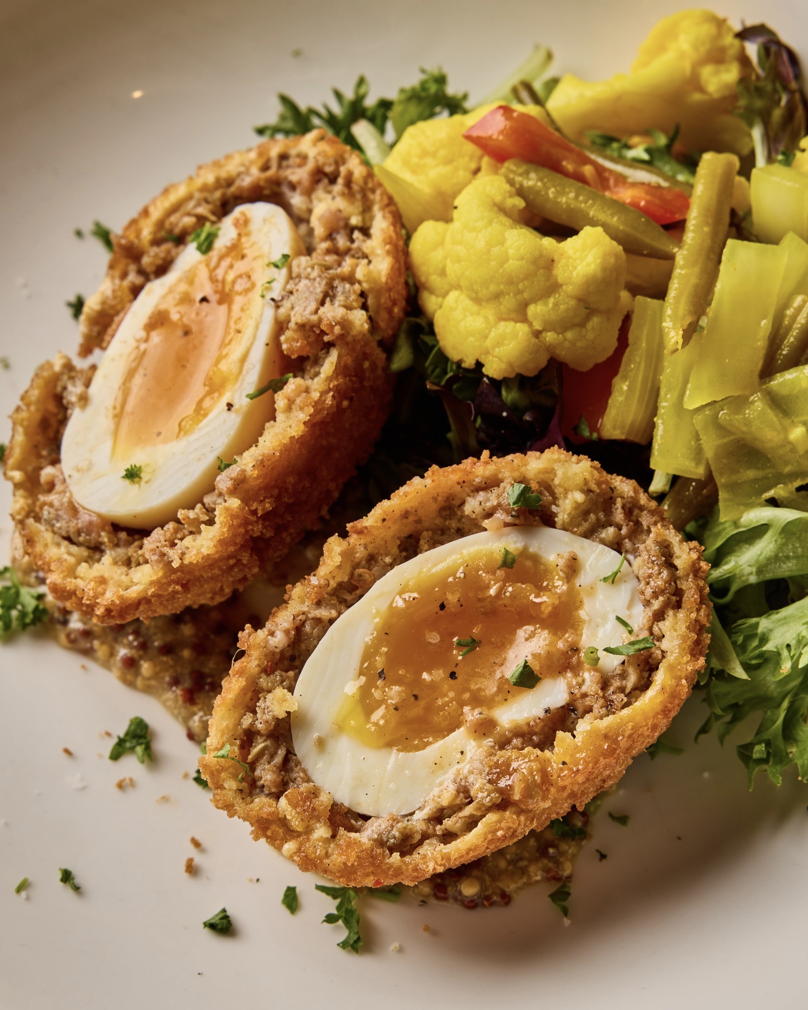 Halved Scotch eggs with runny yolks, served with pickled vegetables and greens.