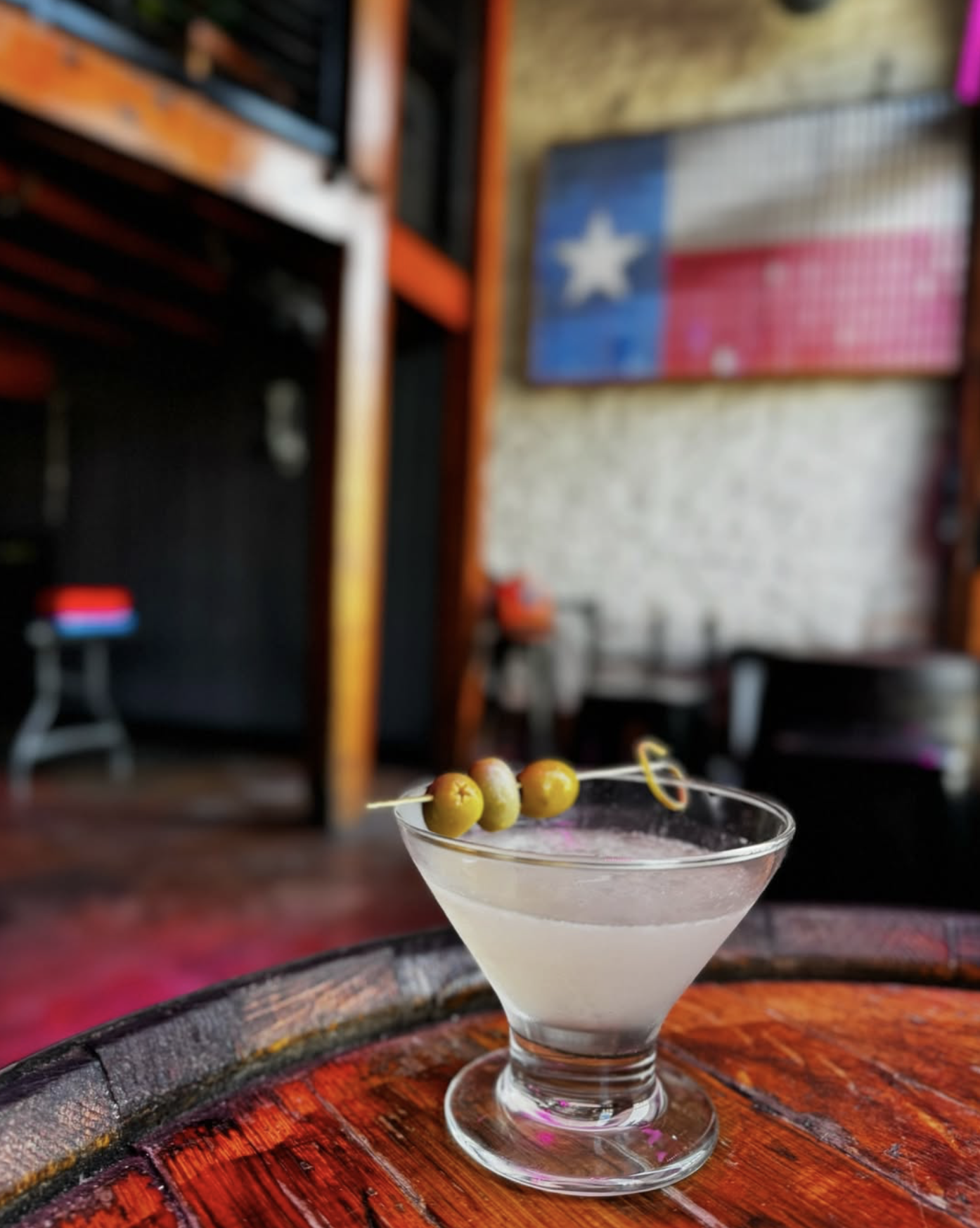 Martini with olives on a wooden barrel, Texas flag in background.