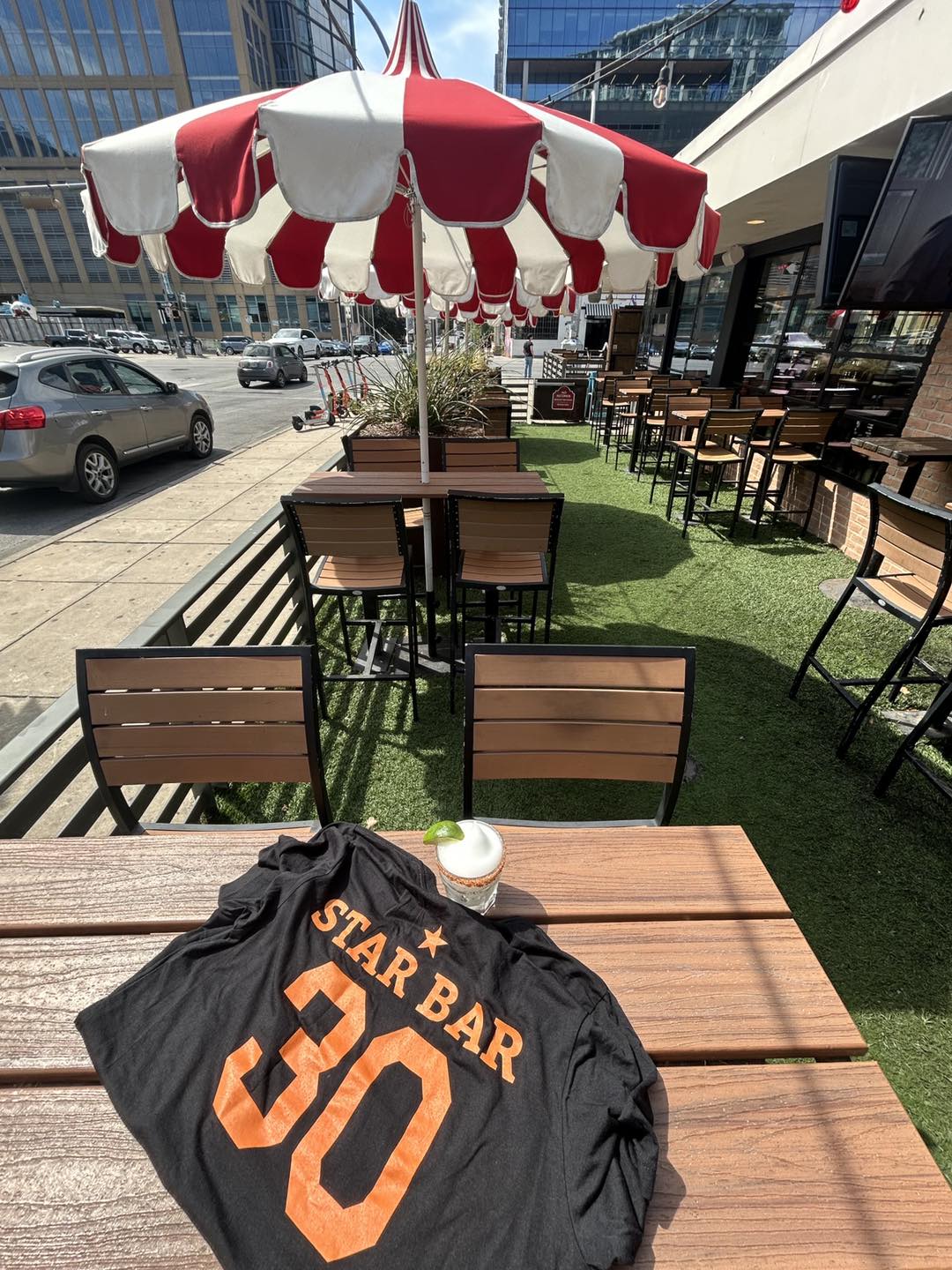 Black Star Bar 30 shirt, drink, and red-white umbrella on patio.
