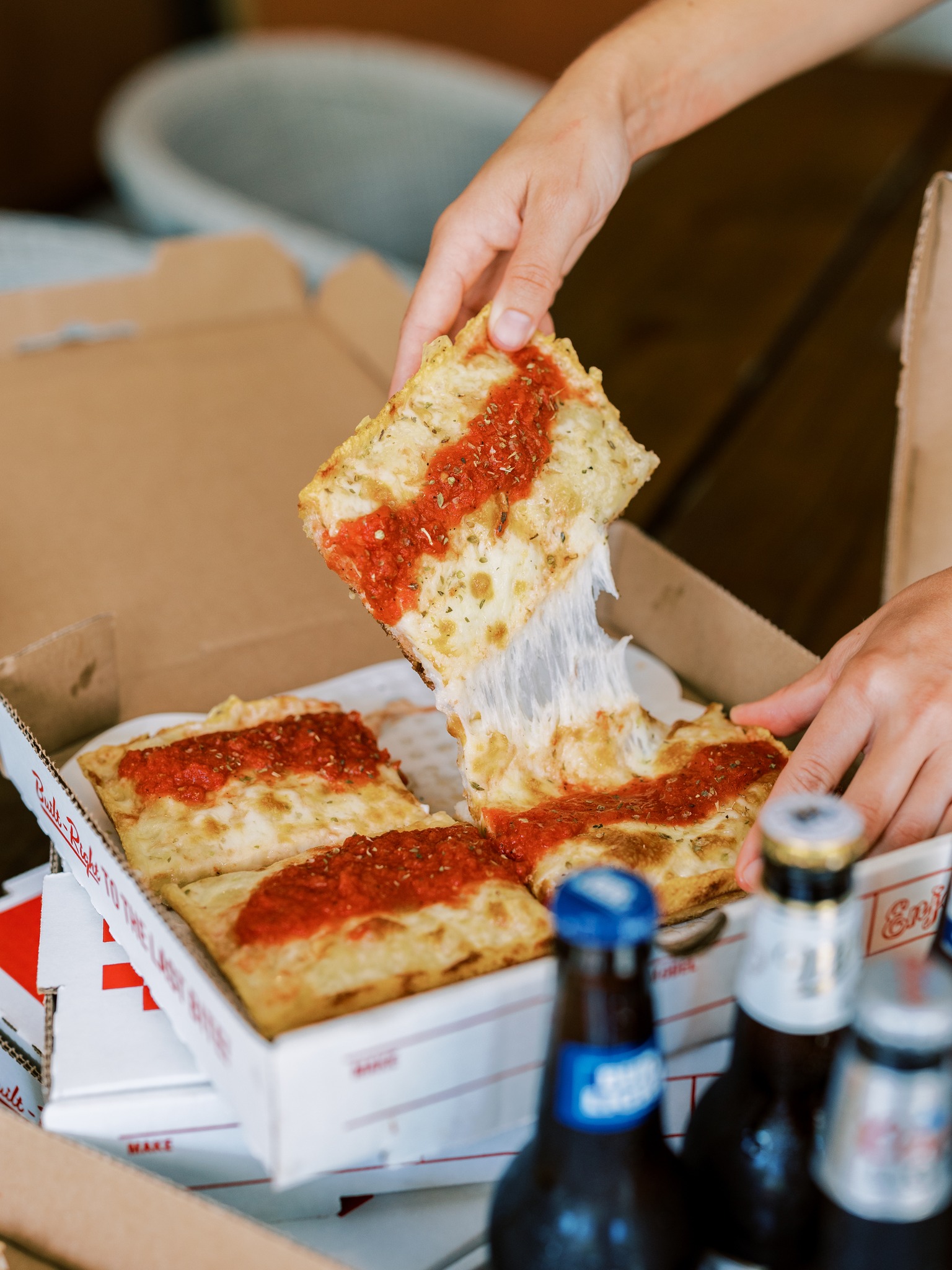 A hand pulls a square pizza slice with stretchy cheese from a box.