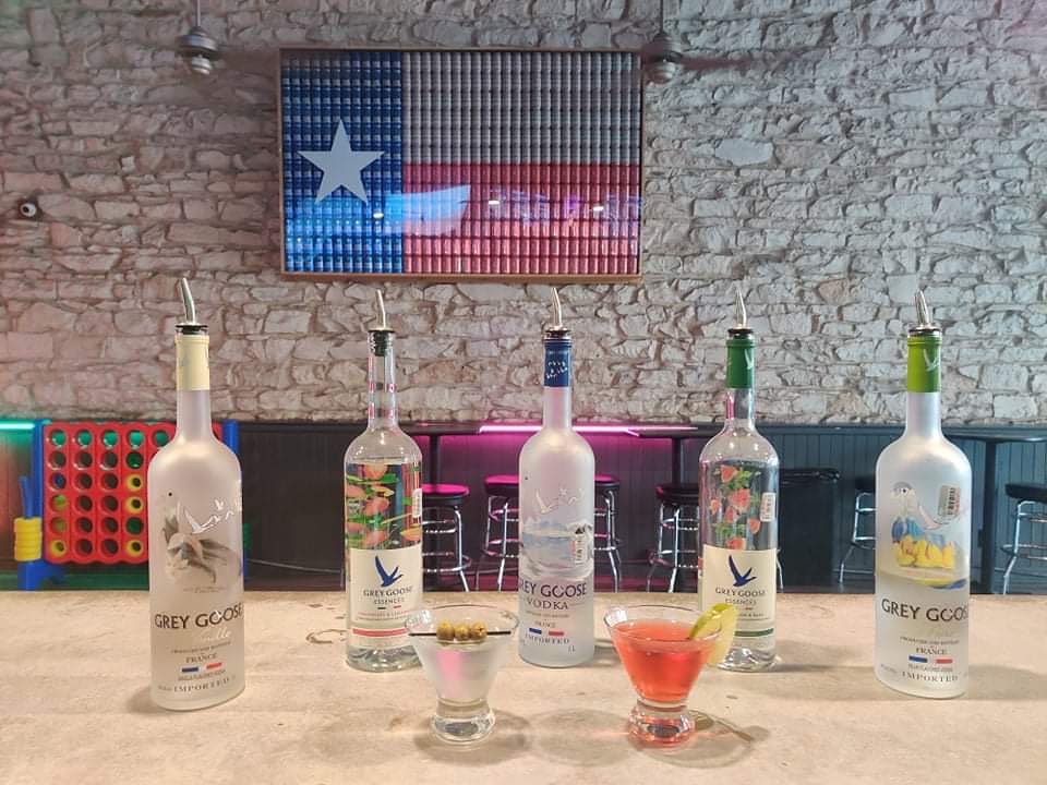 Five Grey Goose vodka bottles, two cocktails, and a Texas flag.