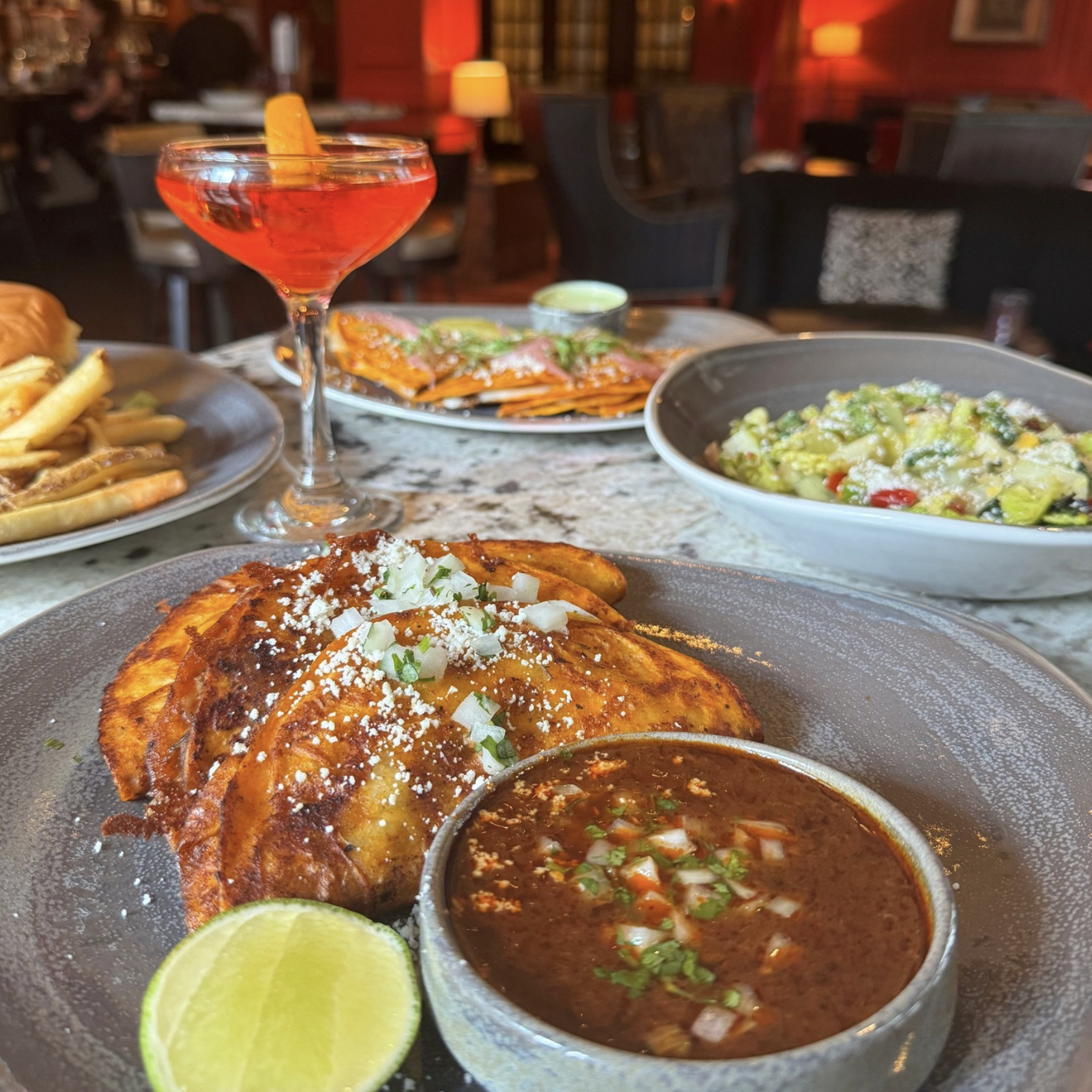 Close-up of crispy tacos, consommé, and a red cocktail at a restaurant.