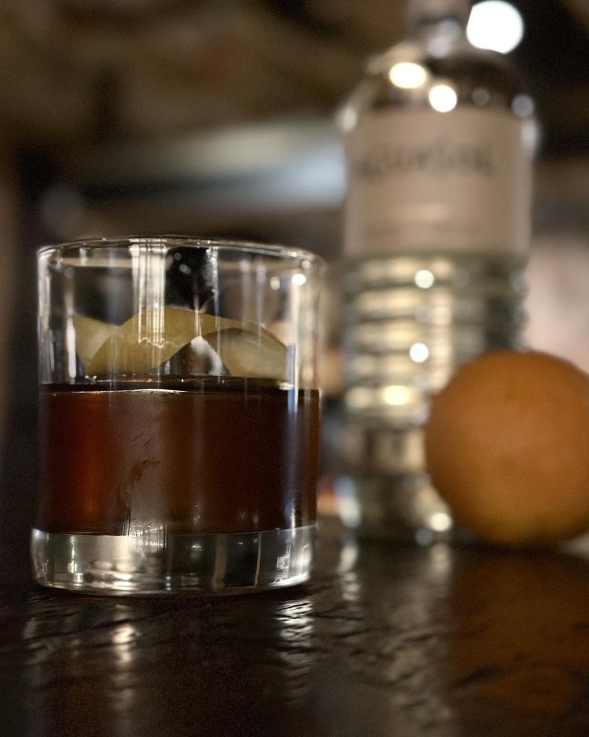 Glass of dark liquid with ice, citrus peel, bottle, and orange.