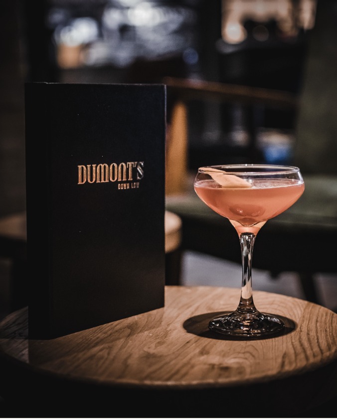 Pink cocktail and Dumont's menu on a wooden table.