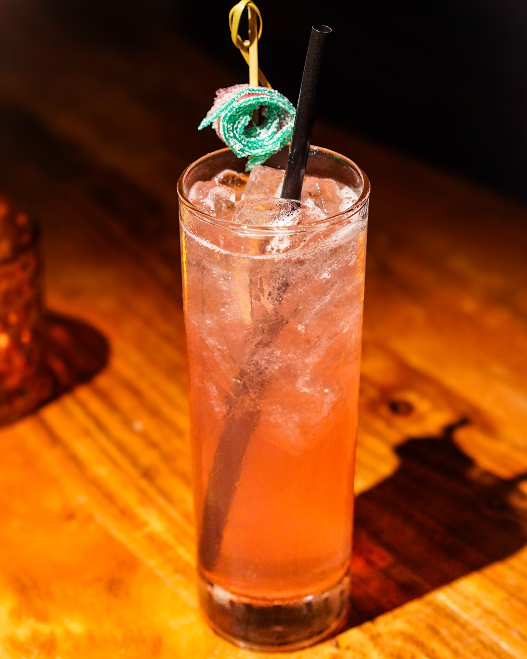 Pink cocktail with ice, straw, and green candy garnish on wood.