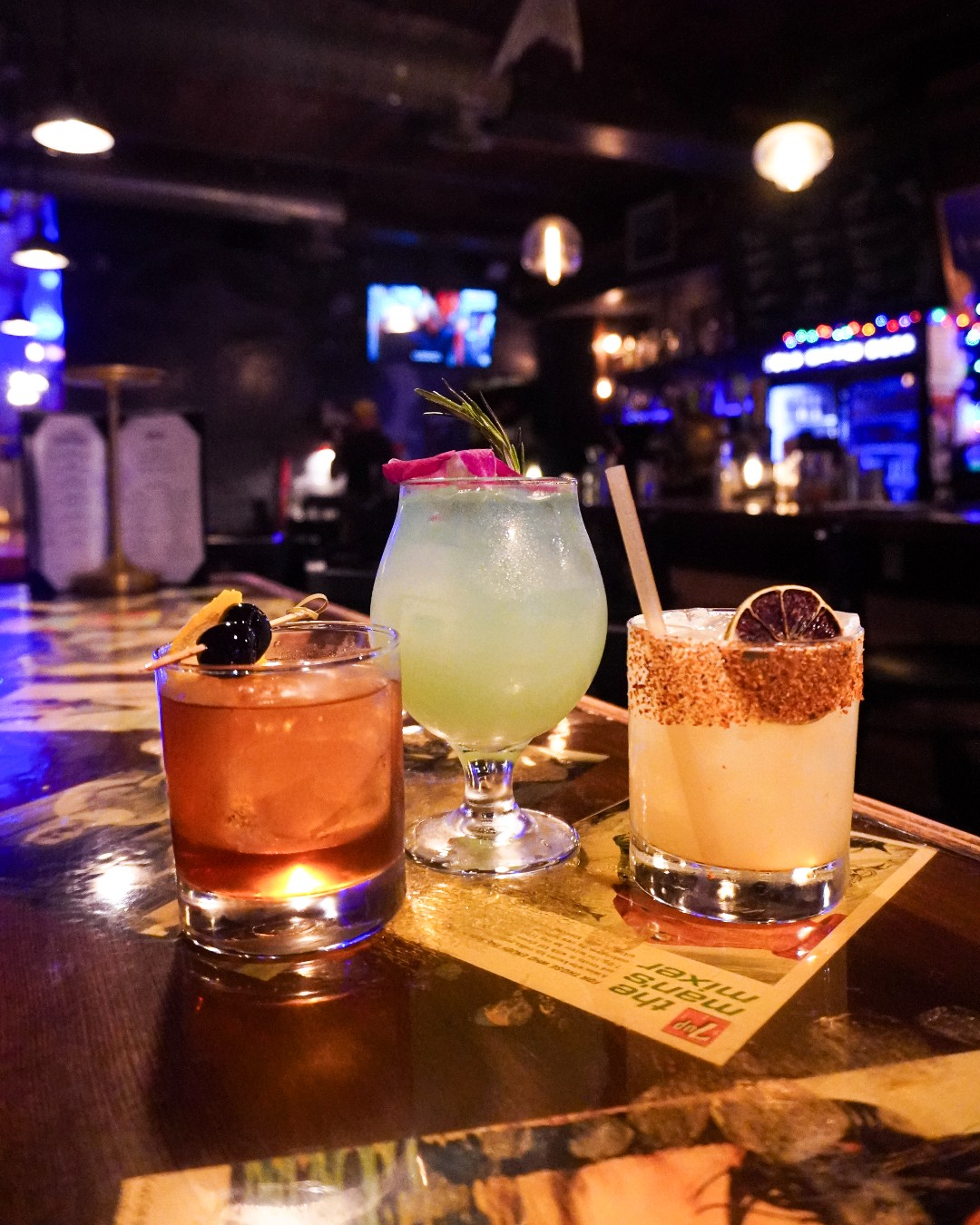 Three garnished cocktails on a bar counter in a dimly lit bar.