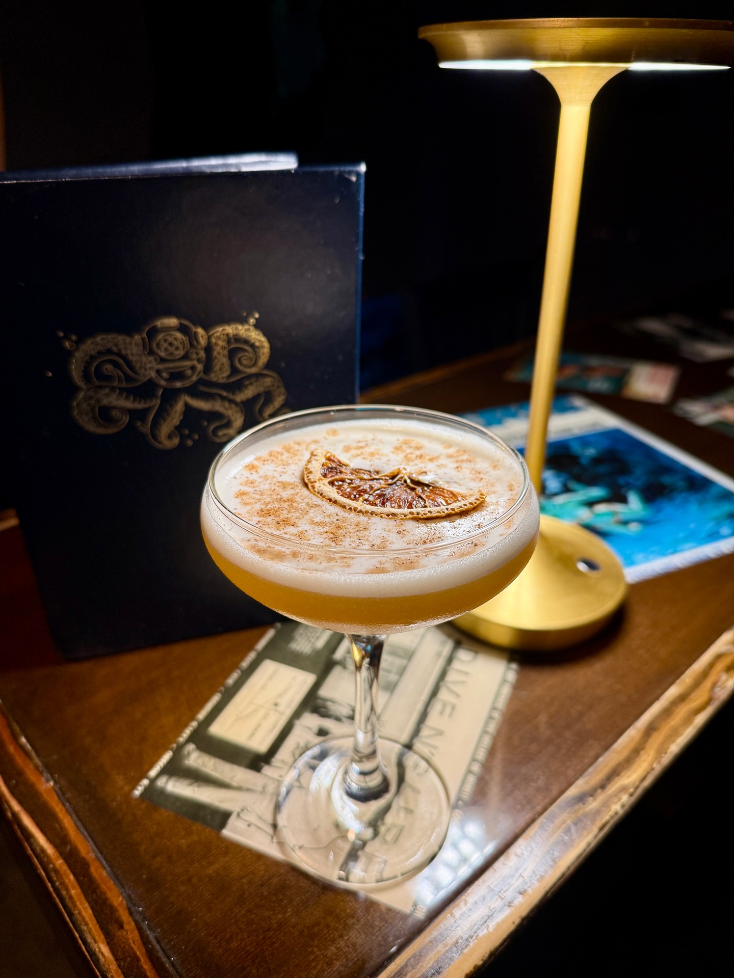 Foamy cocktail with dried orange slice, octopus menu, and gold lamp.