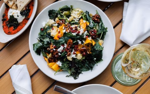 Salad, burrata cheese dish, and white wine on wooden table.