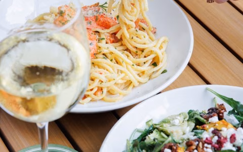 Glass of white wine, pasta, and salad on a wooden table.