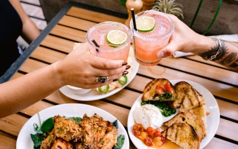 Two hands toast pink cocktails over a table with food.