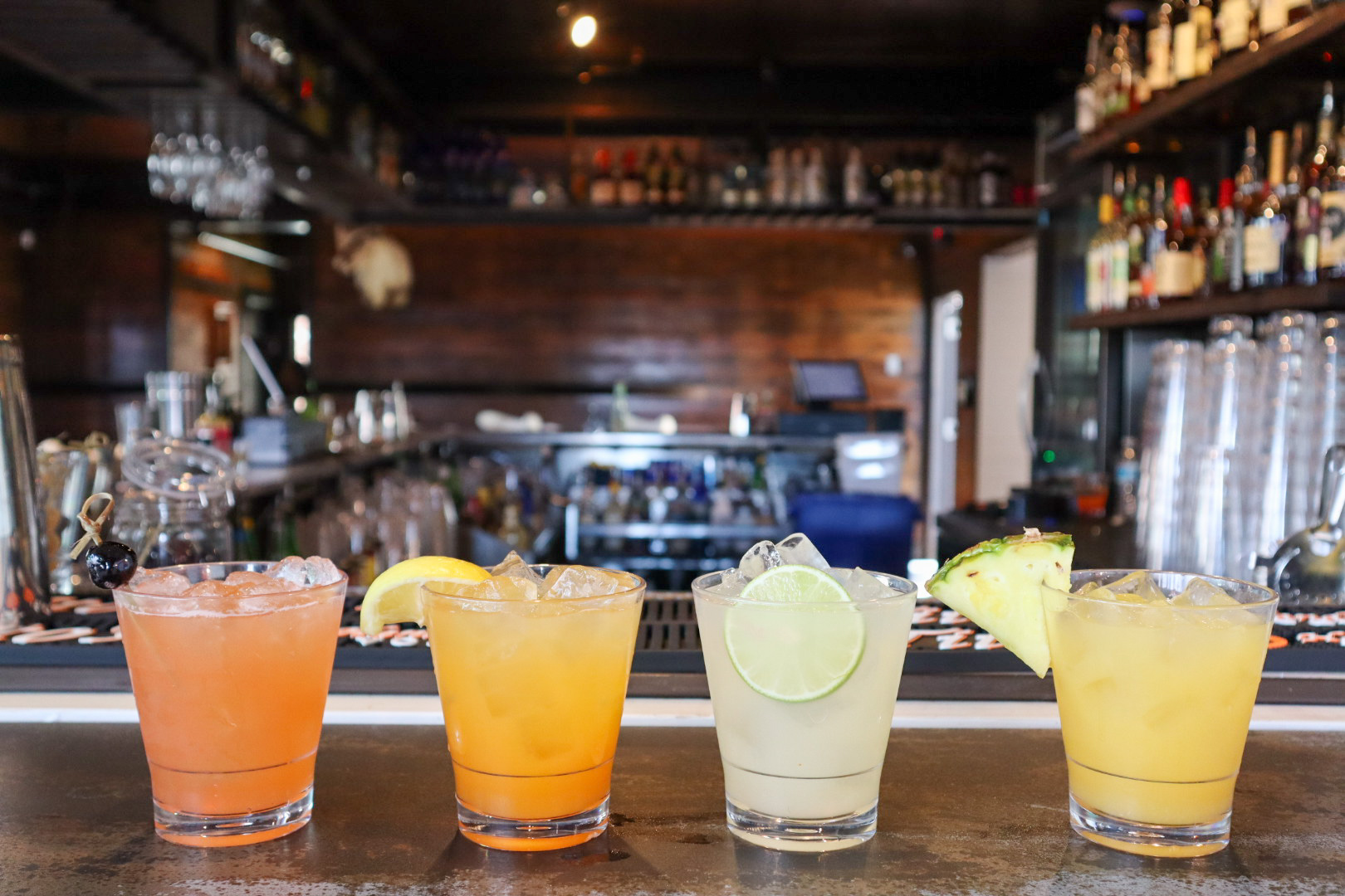 Four colorful cocktails with ice and fruit garnishes on a bar.