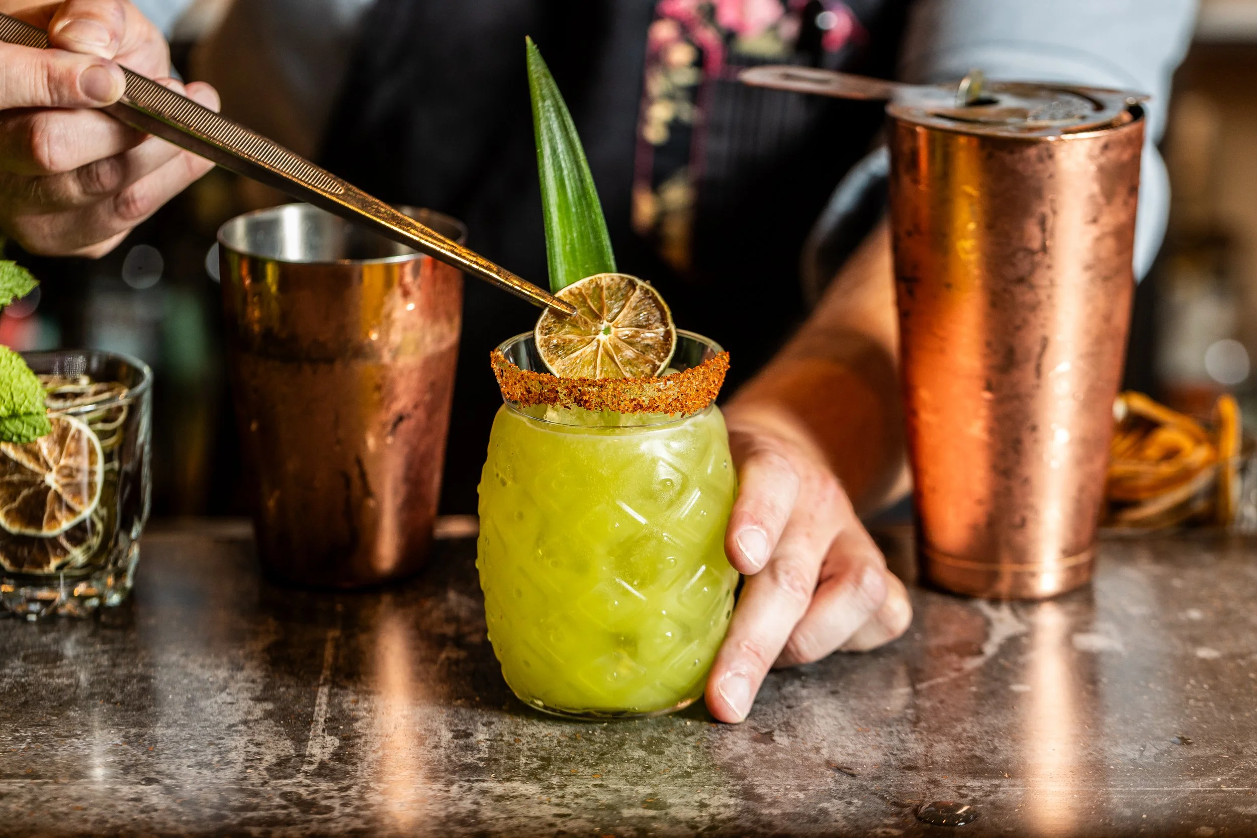 Bartender garnishing a green cocktail with dried lime and tongs.
