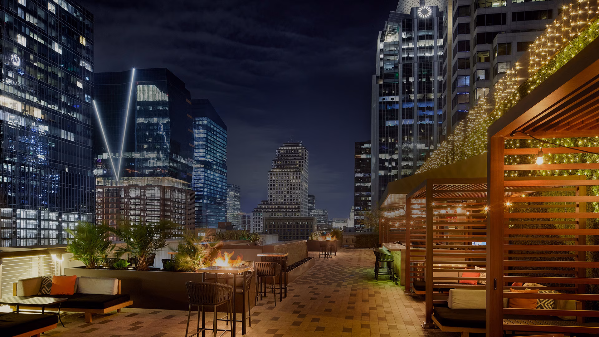 Rooftop patio at night with fire pits, seating, and illuminated city skyline.