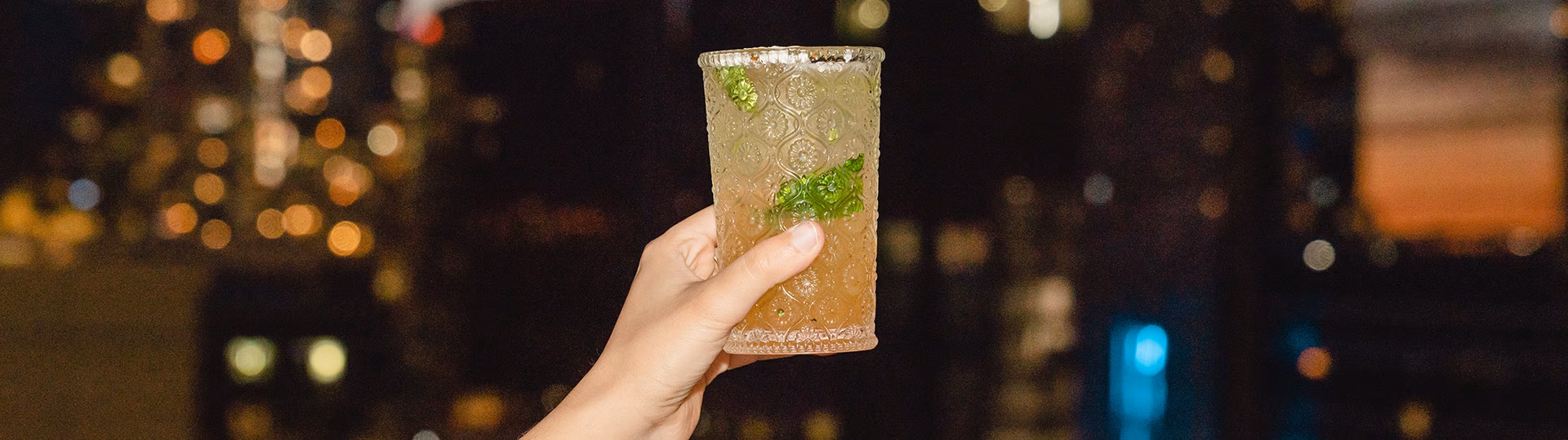 Hand holding a textured glass with mint drink, blurred night city.