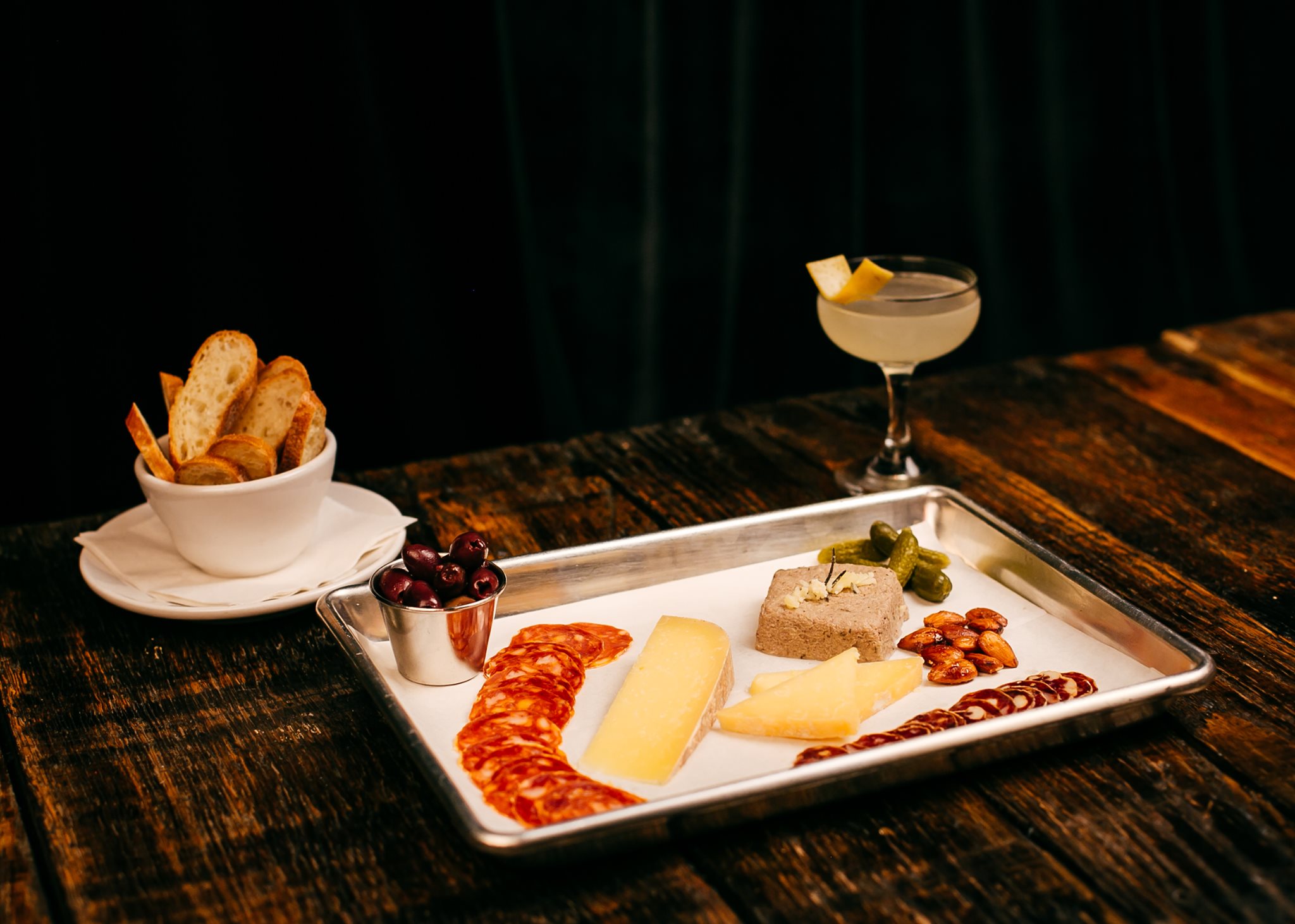 Charcuterie board with cheeses, meats, olives, pickles, bread, and cocktail.